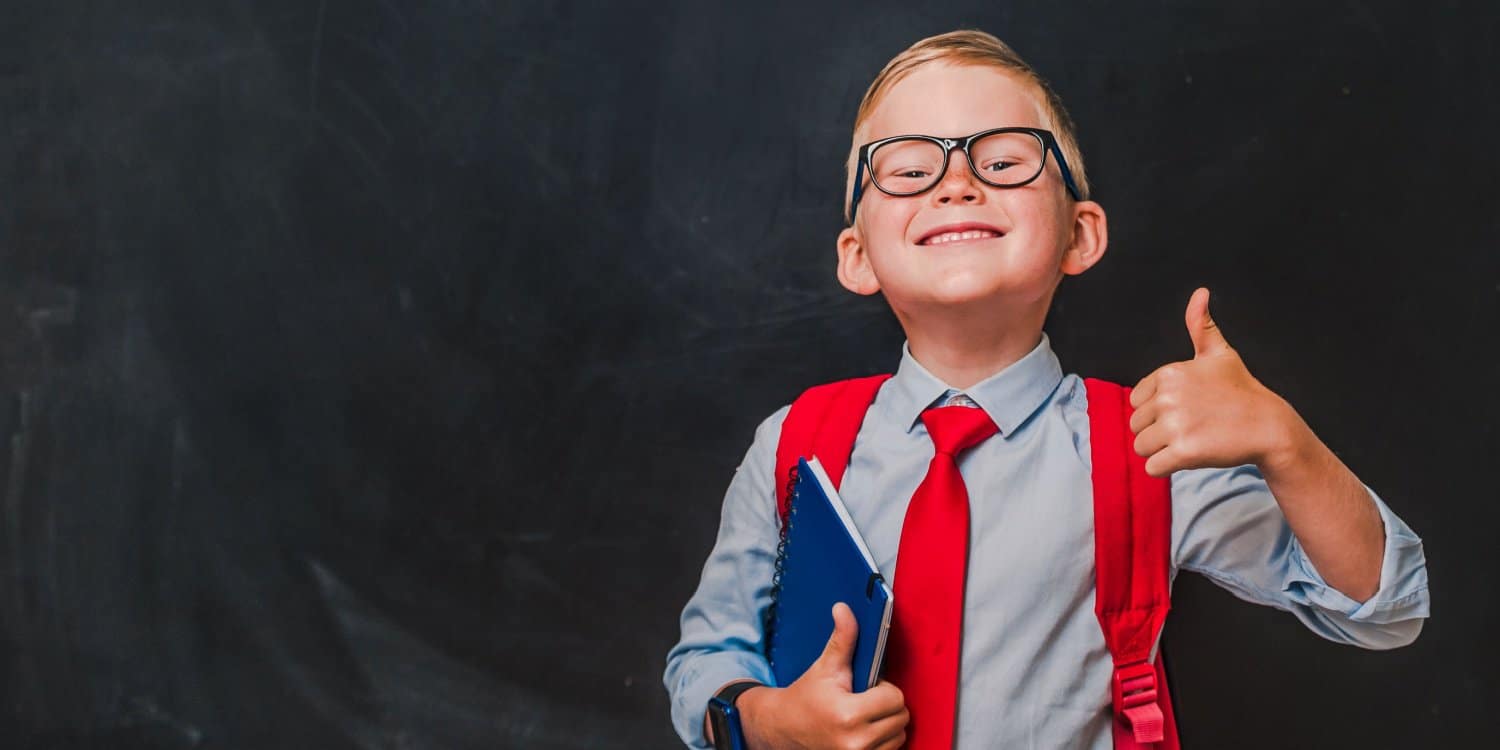 Happy schoolboy in glasses and school uniform standing in front of a blackboard showing a thumbs up sign, having just set up his own limited company.