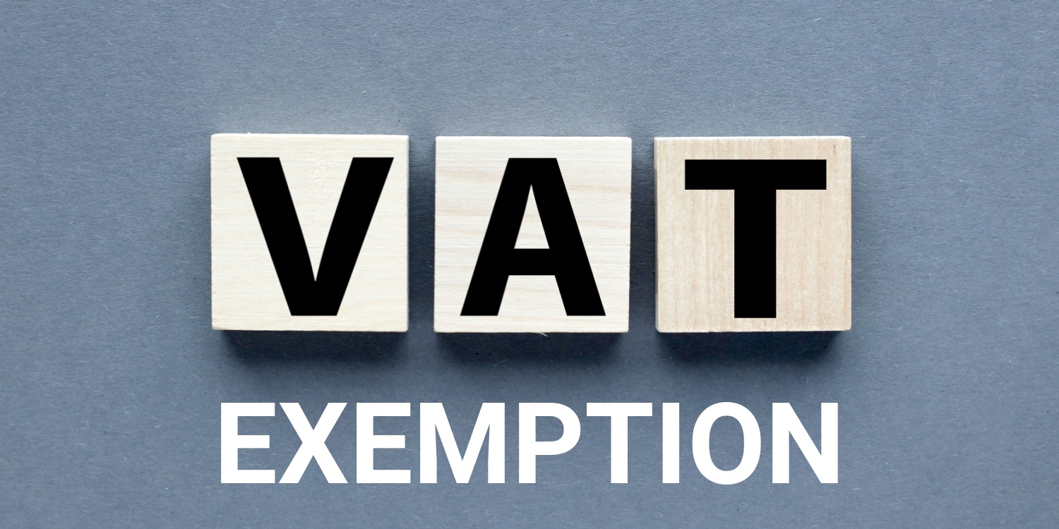 Three wooden cubes on a gray background spelling the word 'VAT' and a heading below in white block capitals of 'EXEMPTION'.