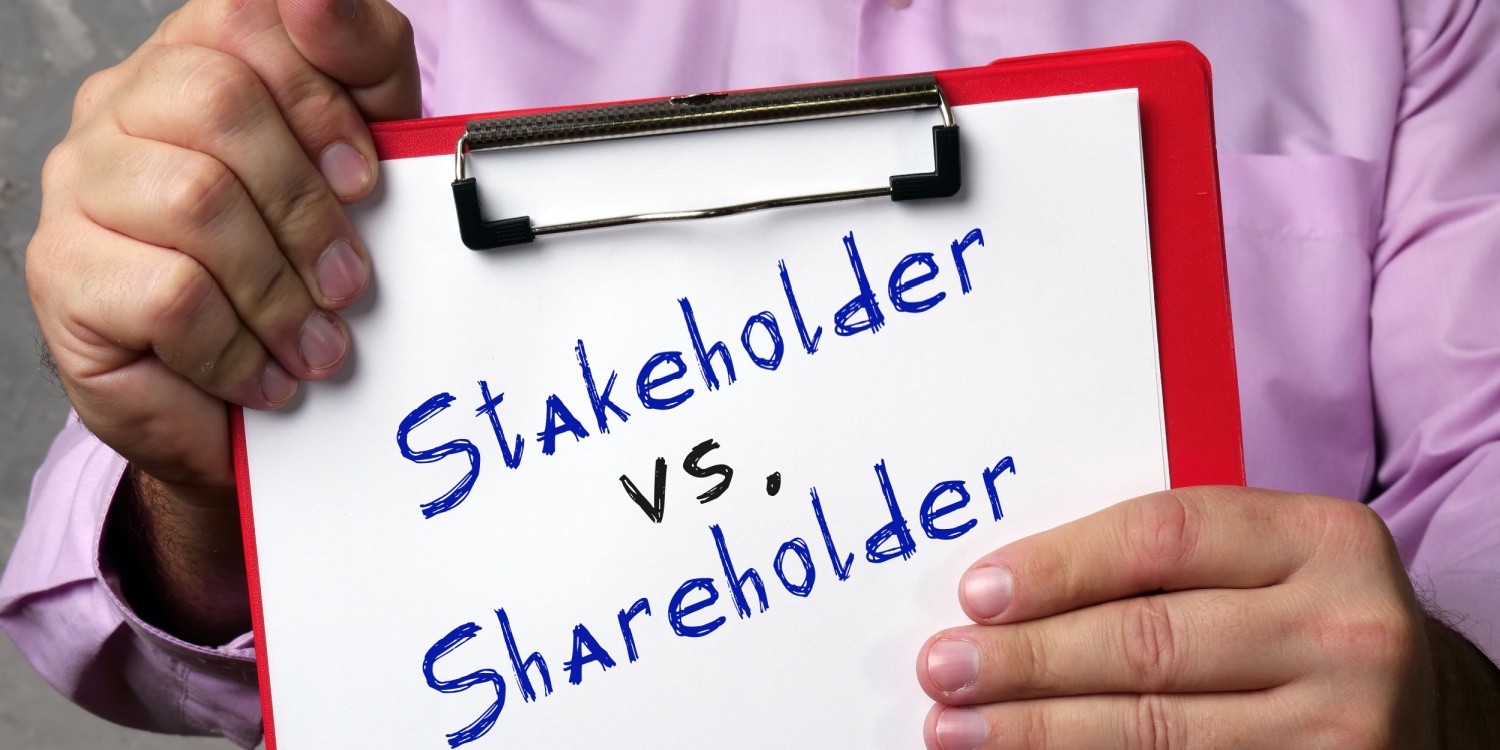 Businessman wearing a pink shirt and holding a clipboard with Stakeholder vs. Shareholder handwritten on a white sheet of paper.