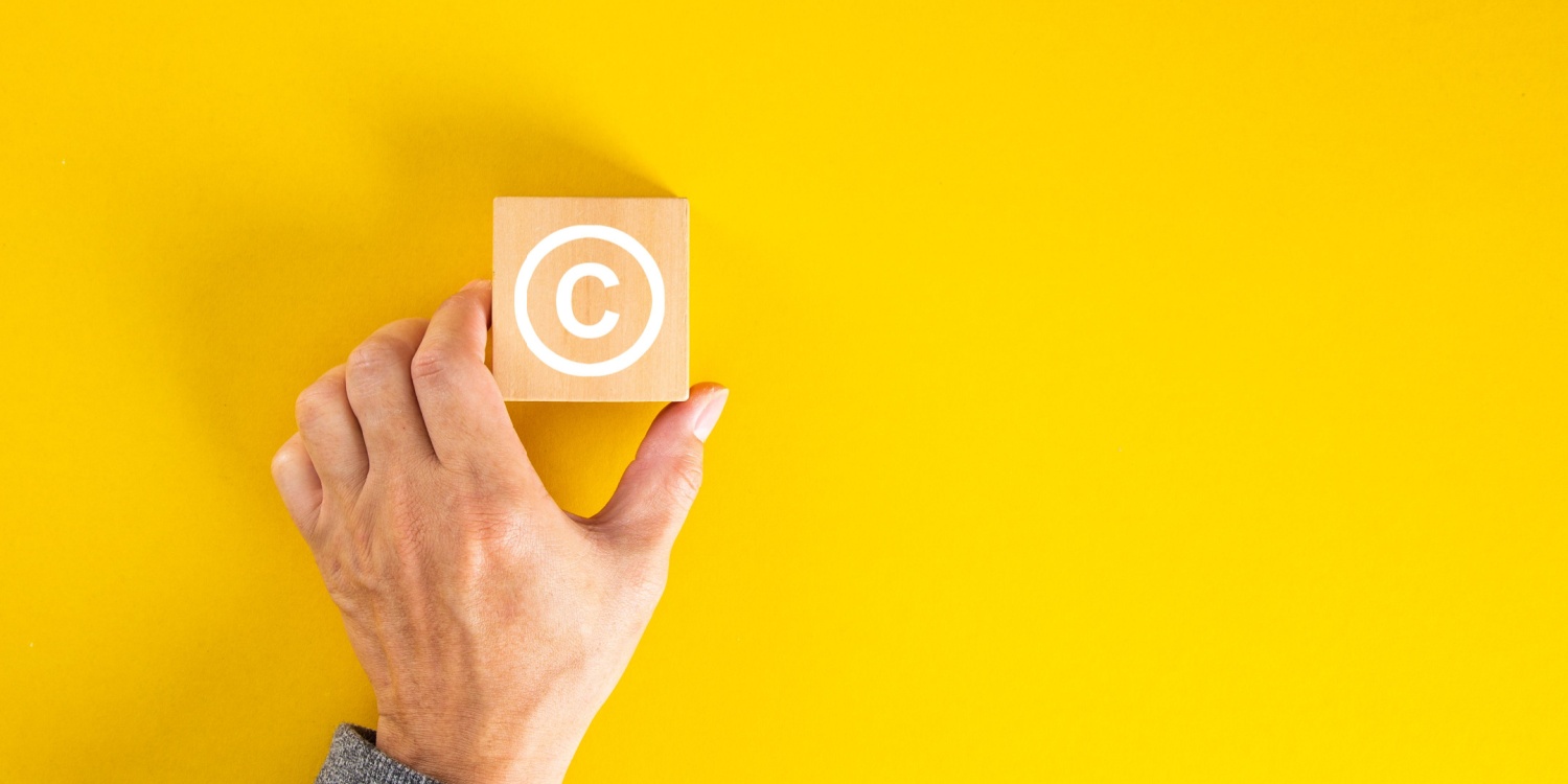 A male hand holding a wooden block with the copyright symbol displayed on the block in white, against a yellow background.