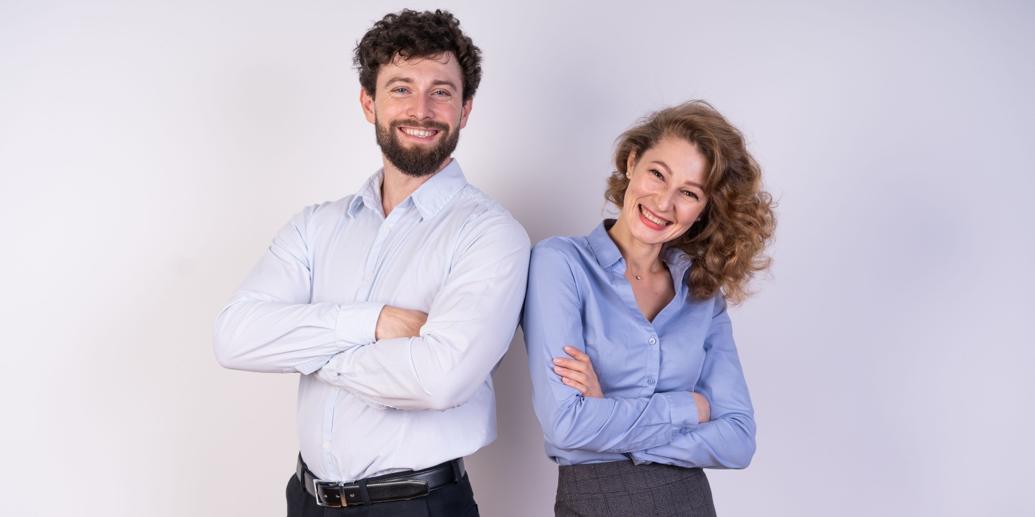 Two new business partners, a male and a female, looking directly at the camera, standing side by side, smiling and cheerful.