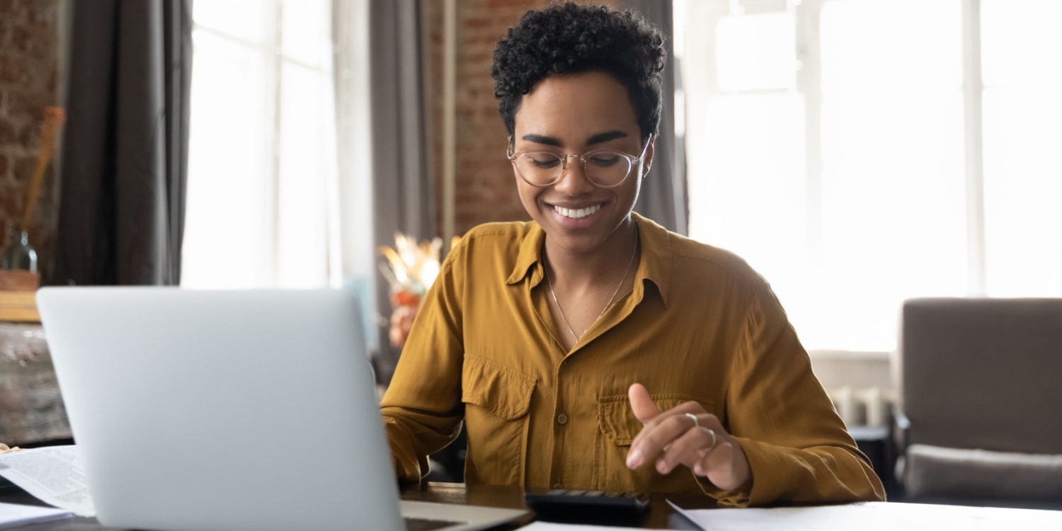 Happy young female entrepreneur working at a desk with a calculator and laptop, using HMRC's new VAT Registration Estimator tool.