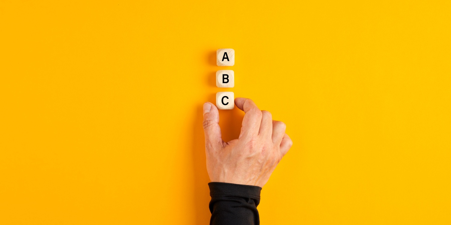 A male hand placing 3 wooden blocks on a yellow table, with the letters A, B and C printed on them, illustrating the concept of alphabet shares.